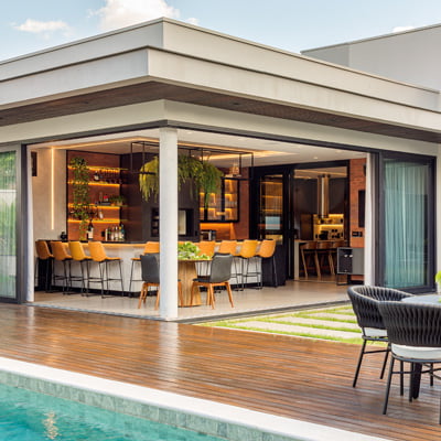 Ambiente aconchegante a beira da piscina, com estrutura coberta que conta com uma grande bancada de 12 lugares, junto de uma churrasqueira. A decoração é moderna e autoral.